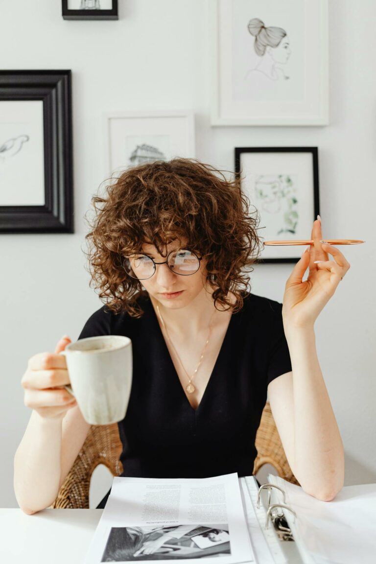 Young woman wearing glasses, reading documents with a coffee mug in a modern art-filled room.